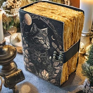 May include: A leather-bound journal with a black cover featuring a detailed illustration of a cat, moon, and floral elements. The pages have a vintage, aged appearance. A black leather strap with a metal clasp secures the journal.