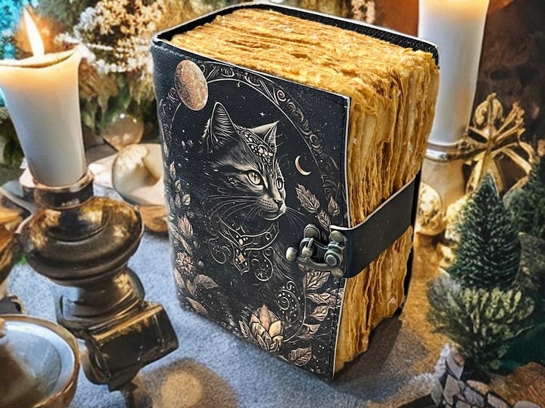 May include: A leather-bound journal with a black cover featuring a detailed illustration of a cat, moon, and floral elements. The pages have a vintage, aged appearance. A black leather strap with a metal clasp secures the journal.
