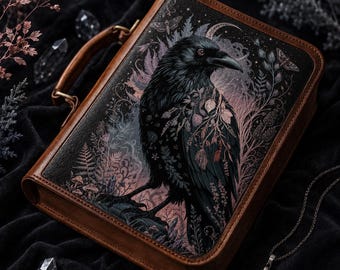 May include: A rectangular brown leather carrying case featuring a detailed illustration of a black raven surrounded by botanical elements, ferns, and a crescent moon. The case has a sturdy top handle and a smooth, structured frame with dark edges.