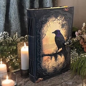 May include: A decorative book with a raven illustration on the cover. The book has a black cover with ornate detailing and a metal clasp. The raven is perched on a branch against a warm, glowing background. Lit candles surround the book, creating a mystical atmosphere.