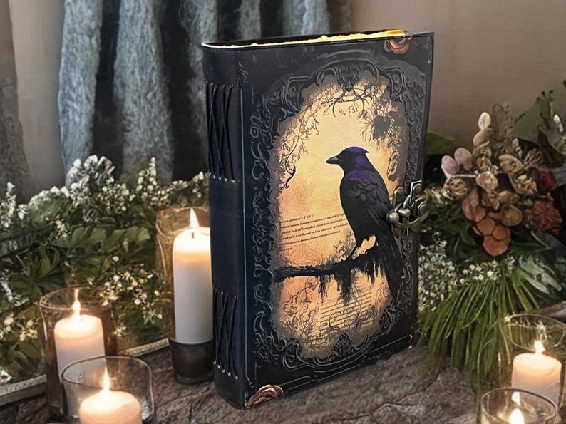 May include: A decorative book with a raven illustration on the cover. The book has a black cover with ornate detailing and a metal clasp. The raven is perched on a branch against a warm, glowing background. Lit candles surround the book, creating a mystical atmosphere.