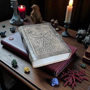 May include: A leather-bound journal with an embossed design featuring a central figure and intricate patterns. The journal rests atop a burgundy leather notebook. A lit candle and decorative items are in the background.