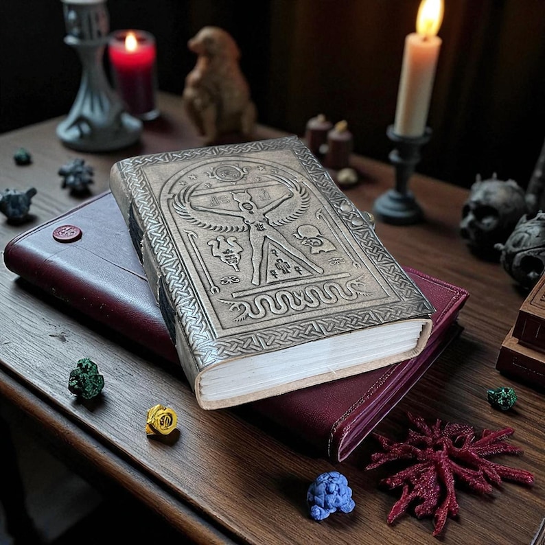 May include: A leather-bound journal with an embossed design featuring a central figure and intricate patterns. The journal rests atop a burgundy leather notebook. A lit candle and decorative items are in the background.