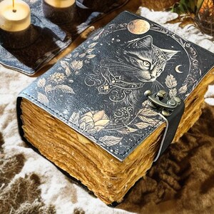 May include: A vintage-style journal with a cat illustration on the cover. The cover is dark blue with gold floral accents and a crescent moon. The journal has aged, yellowed pages and a clasp closure. Candles and decorative elements are in the background.