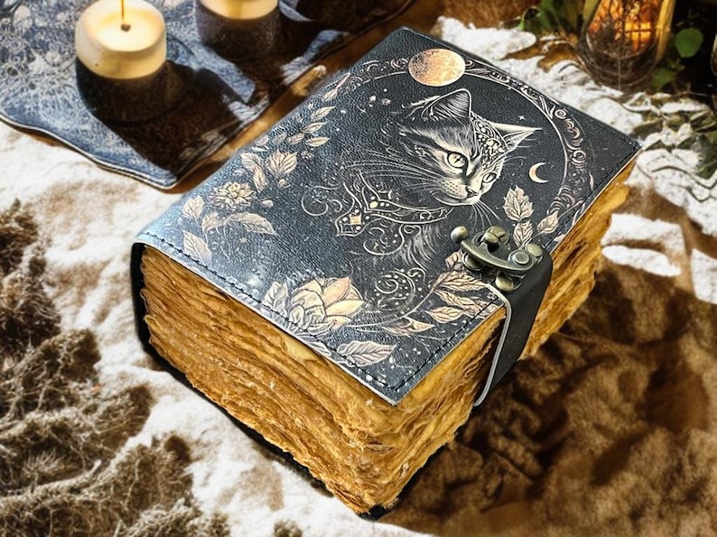 May include: A vintage-style journal with a cat illustration on the cover. The cover is dark blue with gold floral accents and a crescent moon. The journal has aged, yellowed pages and a clasp closure. Candles and decorative elements are in the background.