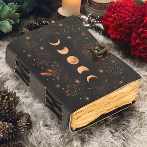 May include: Black leather journal with a moon phase design in gold. The journal has a metal clasp and aged-looking pages. The cover also features botanical and star accents. The journal is closed and resting on a soft, gray surface.