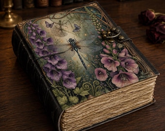May include: A hardcover journal with a dark, textured cover featuring a central dragonfly illustration surrounded by purple foxglove flowers and small butterflies. The book includes a brass-colored metal latch closure and thick, deckled-edge paper pages.