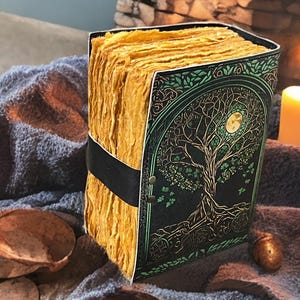 Puede incluir: Un diario hecho a mano con un dise&ntilde;o de &aacute;rbol de la vida en la portada. La portada es negra con detalles verdes y una luna llena. Las p&aacute;ginas son gruesas y amarillentas, sujetas con una correa negra. Una vela encendida est&aacute; en el fondo.