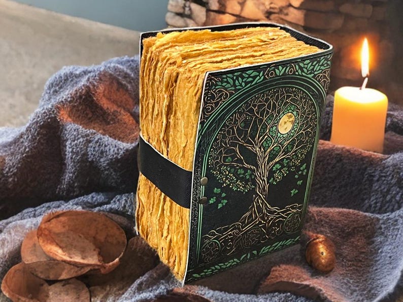 Puede incluir: Un diario hecho a mano con un dise&ntilde;o de &aacute;rbol de la vida en la portada. La portada es negra con detalles verdes y una luna llena. Las p&aacute;ginas son gruesas y amarillentas, sujetas con una correa negra. Una vela encendida est&aacute; en el fondo.