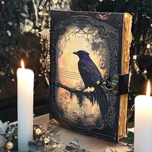 May include: An antique-style journal with a raven illustration on the cover. The cover is dark with an ornate frame and a clasp. The pages have a vintage, aged appearance. Two white candles are lit on either side of the journal.