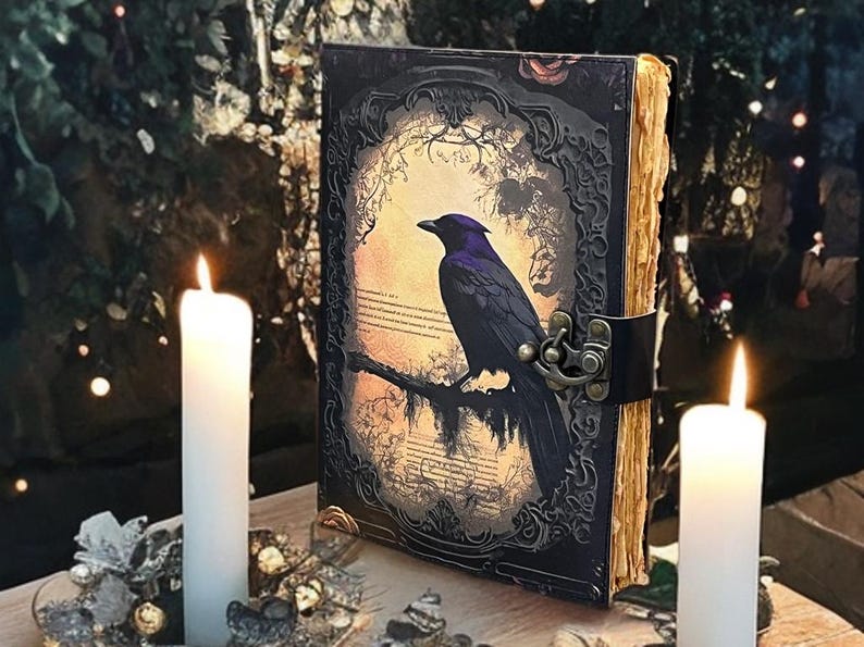 May include: An antique-style journal with a raven illustration on the cover. The cover is dark with an ornate frame and a clasp. The pages have a vintage, aged appearance. Two white candles are lit on either side of the journal.