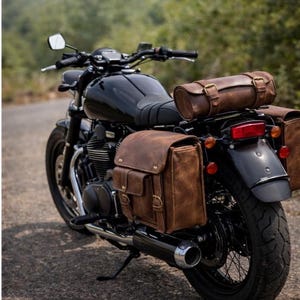 May include: Black motorcycle with brown leather saddlebags and a matching roll bag. The motorcycle is parked on a paved road with a blurred green background. The leather bags have brass buckles.