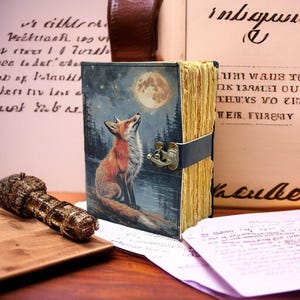 Könnte beinhalten: Ein ledergebundenes Notizbuch mit einer Illustration von Fuchs und Mond. Das Cover zeigt einen Fuchs, der einen Vollmond über einem Wald betrachtet. Das Notizbuch hat einen Verschluss und gealterte Seiten. Ein dekorativer Stift und Papiere sind ebenfalls sichtbar.