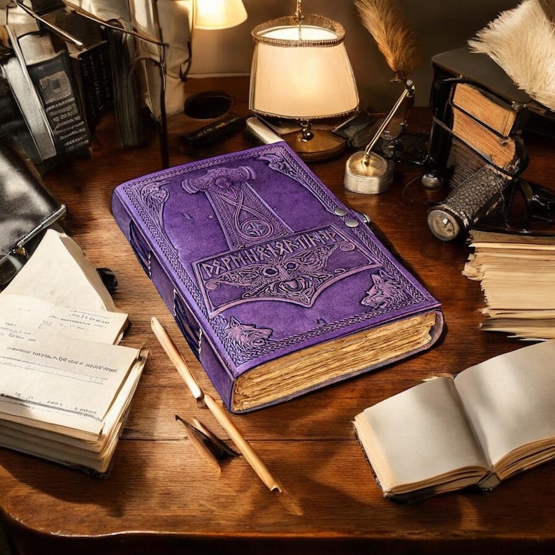 May include: A purple leather-bound journal with embossed Viking-style artwork, including a hammer and wolf designs. The journal is open, revealing aged paper pages. The cover features intricate details and a clasp closure. The journal sits on a wooden desk.