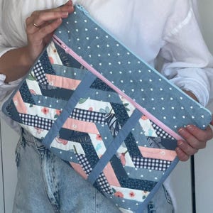 Puede incluir: Un bolso de mano acolchado con exterior de mezclilla azul y cremallera rosa. El bolso presenta un diseño de patchwork con piezas de tela floral, cuadros y lisas en tonos de azul, rosa y blanco. La sección superior tiene un fondo azul claro con corazones blancos.