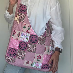 Könnte beinhalten: Eine rosa Patchwork-Umhängetasche mit einem sich wiederholenden Smiley-Gesicht-Design. Die Tasche hat ein Blumen- und geometrisches Muster sowie einen Quastenbesatz. Die Tasche ist aus Stoff.
