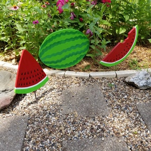 May include: Three wooden watermelon decorations, painted in red and green, with black seeds. The watermelon slices are on sticks, and the whole watermelon is flat. The decorations are on a bed of gravel.
