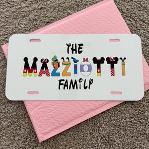 May include: White license plate with the text "THE MAZZIOTTI FAMILY" in black and colorful Disney-themed lettering. The plate is on top of a pink padded envelope.