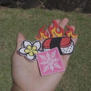 Puede incluir: Un conjunto de tres parches bordados que incluye una flor de plumeria blanca y amarilla, una pieza de sushi coronada con llamas rojas y amarillas, y un parche cuadrado rosa con un diseño de copo de nieve blanco con detalles ramificados.