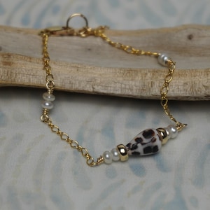 May include: A gold-colored chain bracelet featuring a central, speckled bead in white and brown, flanked by small, white pearls and gold-colored beads. The bracelet is displayed on a piece of driftwood.