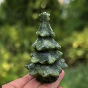 May include: A small, carved green stone Christmas tree. The tree has a star on top and layered branches. The stone has a mottled appearance with variations in the green color. The tree is held in a hand against a blurred green background.