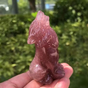 Puede incluir: Una figura de cristal tallada, de color púrpura rojizo, de un lobo en posición sentada. El lobo está detallado con pelaje y rasgos faciales visibles. El fondo es follaje verde borroso.