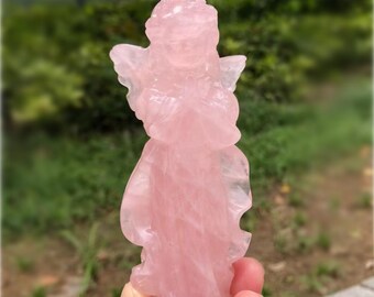 May include: A carved rose quartz angel figurine. The angel has folded hands and wings, and is a soft pink color. The figurine is held in a person's hand against a blurred green background.