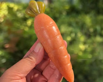 May include: A realistic orange carrot sculpture with a green stem. The carrot is held in a hand, showcasing its smooth texture and detailed ridges. The background is a blurred green, suggesting an outdoor setting.