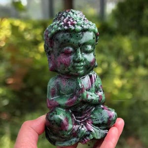 May include: A small Buddha statue in a seated pose, carved from a green and maroon stone. The figure has its hands clasped in prayer. The stone has a speckled appearance, with the colours blending throughout the sculpture.