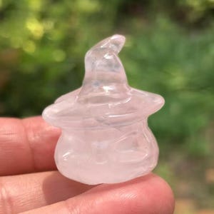 Puede incluir: Una talla de cristal rosa claro translúcido de un sombrero de bruja. El sombrero tiene una base redondeada y una parte superior puntiaguda y curvada. El cristal es liso y parece pulido, con sutiles patrones internos. El fondo es verde borroso.