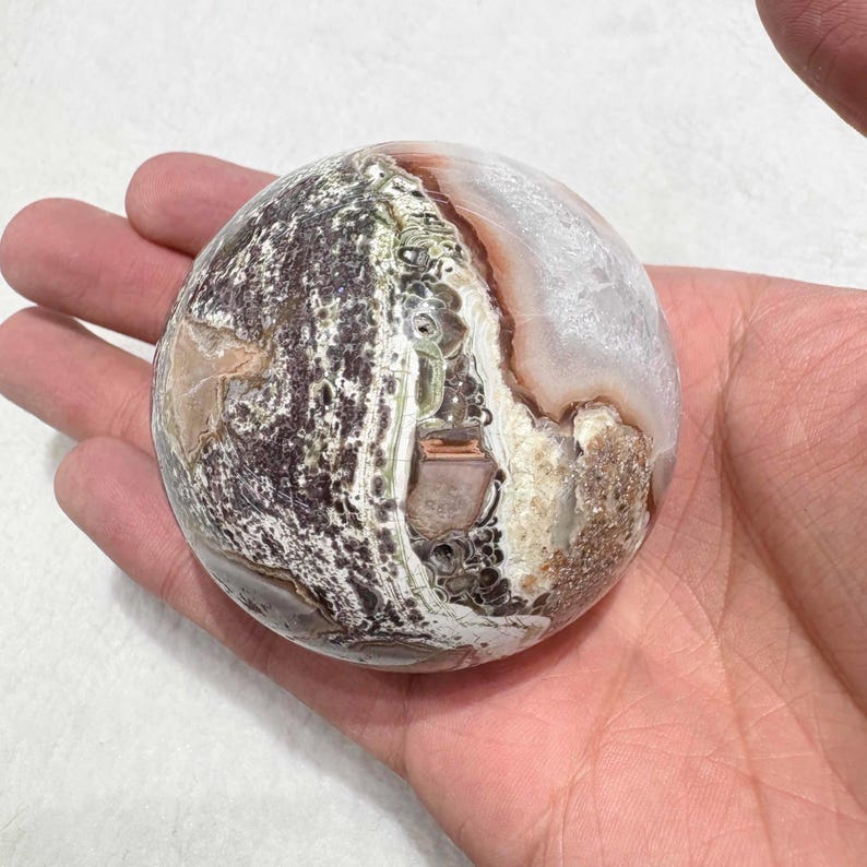 May include: A polished, spherical gemstone with a complex pattern of colors. The stone features areas of brown, white, and green, with a marbled appearance. The sphere is held in a hand, showcasing its size and texture. This decorative stone is a unique home decor item.