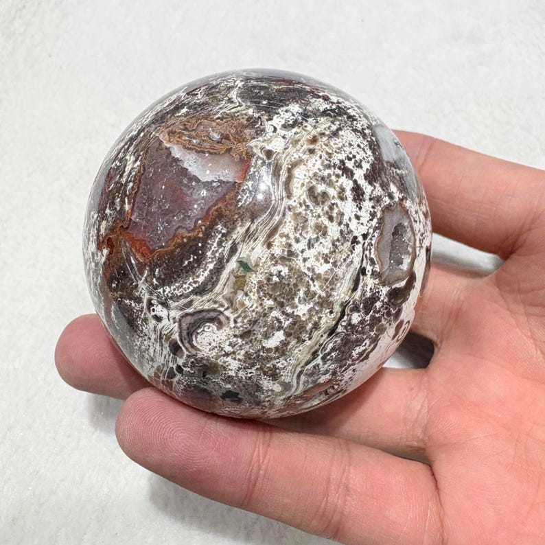 May include: A polished, spherical gemstone with a complex pattern of brown, white, and reddish-purple hues. The orb-shaped stone has a smooth surface and appears to be a decorative item or collectible. The stone is held in a hand.