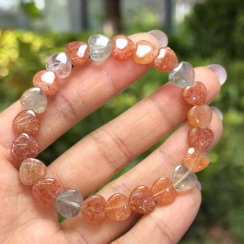 May include: A heart-shaped gemstone bracelet. The bracelet features a variety of translucent, heart-shaped stones in shades of orange, red, and clear. The stones are strung together to form a complete bracelet. The bracelet is held in a hand.