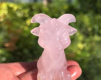 Op de afbeelding: Een roze kwarts geitenbeeldje. De gebeeldhouwde sculptuur toont een gedetailleerde geit met hoorns en een lachend gezicht. Het beeldje wordt in een hand gehouden, met een vage groene achtergrond.