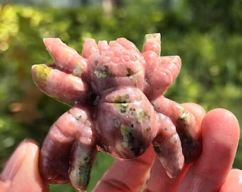 May include: A pink and green carved stone spider figurine. The spider has eight legs and a detailed body. The stone has a natural, textured appearance with variations in color. The figurine is held in a hand against a blurred green background.