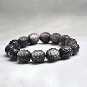 May include: A bracelet composed of round, dark gray beads with a metallic sheen. Each bead features a carved, segmented pattern resembling a pumpkin or a flower, creating a textured surface throughout the circular arrangement of the jewelry piece.
