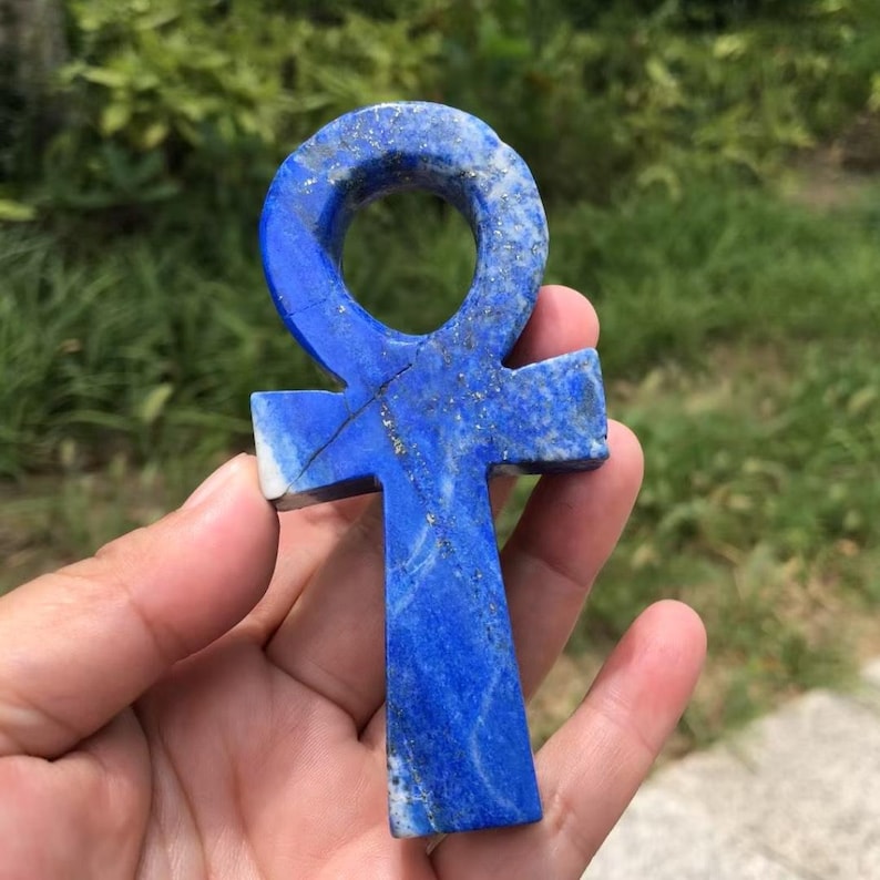 May include: A vibrant blue lapis lazuli ankh pendant. The ankh, a symbol of life, features a loop at the top and a cross-like shape below. The stone has natural gold flecks and a crack. The pendant is held in a hand.