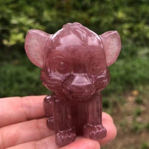 May include: A carved, reddish-purple crystal figurine of a lion cub. The cub is in a seated position with its ears perked up. The crystal has a translucent quality, and the details of the cub's face and body are visible.