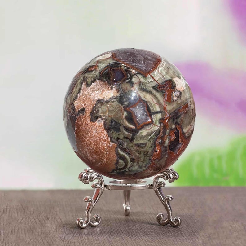 May include: A decorative sphere with a marbled pattern in shades of brown, green, and red, displayed on a silver-colored metal stand. The stand has an ornate, scrolled design. The sphere is a decorative item.
