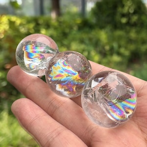 Puede incluir: Tres esferas de cristal de cuarzo transparente con refracciones de luz internas de color arcoíris. Las esferas se sostienen en una mano, con un fondo verde borroso de follaje.