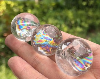 May include: Three clear quartz crystal spheres with internal rainbow-coloured light refractions. The spheres are held in a hand, with a blurred green background of foliage.