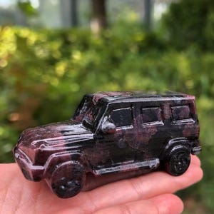 Könnte beinhalten: Ein kleiner, geschnitzter SUV aus Stein in Schwarz- und Burgundertönen. Das Miniaturfahrzeug hat detaillierte Fenster, Räder und einen Kühlergrill. Der Stein hat ein natürliches, strukturiertes Aussehen, und das Auto wird in einer Hand gehalten.