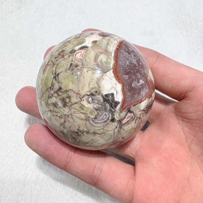 May include: A spherical, polished stone with a mix of green, brown, and white hues. The stone has a cracked, textured surface and a section of purple-red color. The object is held in a hand.