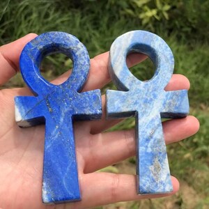 May include: Two lapis lazuli ankh pendants. The pendants are a deep blue color with gold flecks and a cross-like shape with a loop at the top. The ankhs are held in a hand against a blurred green background.