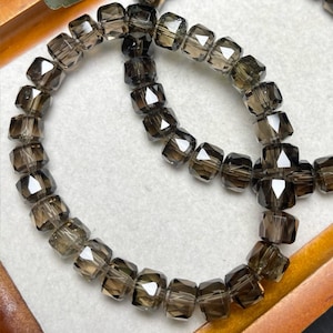 May include: Strands of faceted, smoky quartz beads. The beads are a translucent, brownish-gray color and are strung together. The beads are square-shaped with multiple facets, creating a sparkling effect. The beads are arranged in a circular pattern.
