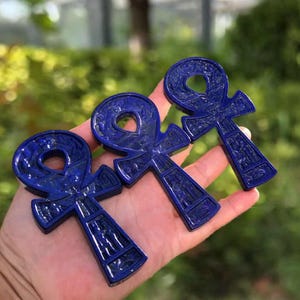 May include: Three dark blue ankh pendants, each with intricate carved details, are displayed. The ankhs are held in a hand against a blurred green background. The pendants have a loop at the top, a vertical bar, and a horizontal bar.