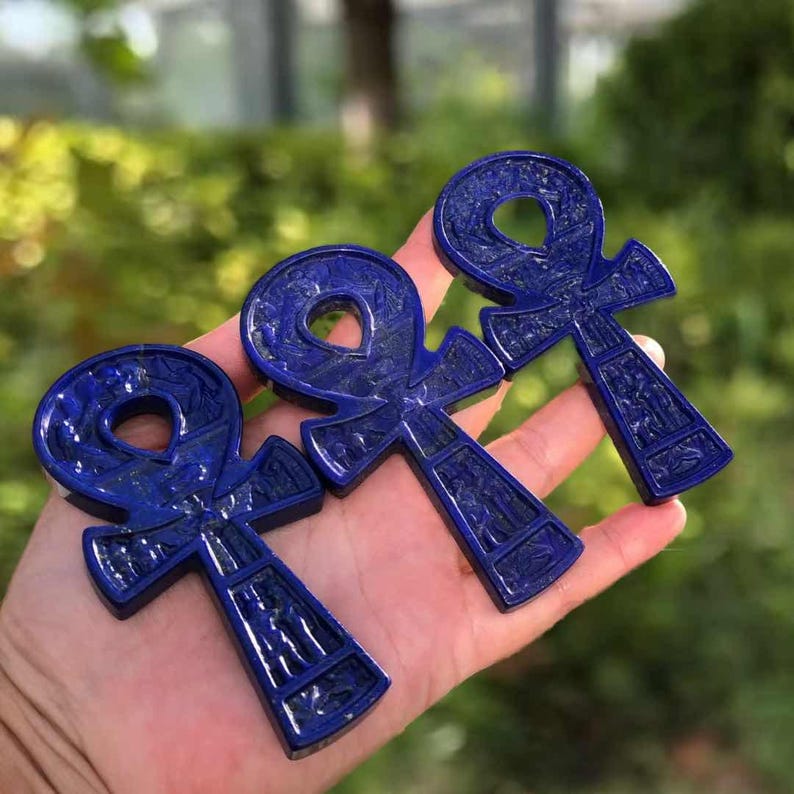 May include: Three dark blue ankh pendants, each with intricate carved details, are displayed. The ankhs are held in a hand against a blurred green background. The pendants have a loop at the top, a vertical bar, and a horizontal bar.