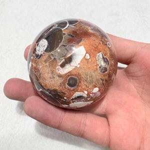 May include: A polished sphere with a unique pattern of brown, tan, and white markings. The orb has a smooth, rounded shape and a glossy finish. The sphere is held in a hand, showcasing its size and texture. The overall theme is decorative objects.