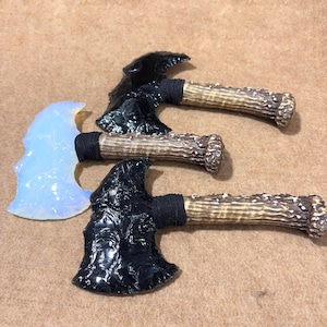May include: Three handcrafted axes with obsidian and opalescent blades. The axe heads are attached to antler handles with black wrapping. The axes are displayed on a brown surface.