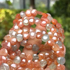May include: Several strands of gemstone bead bracelets. The beads are a mix of orange, clear, and gray colors. The bracelets are held in a hand, with a blurred green background.